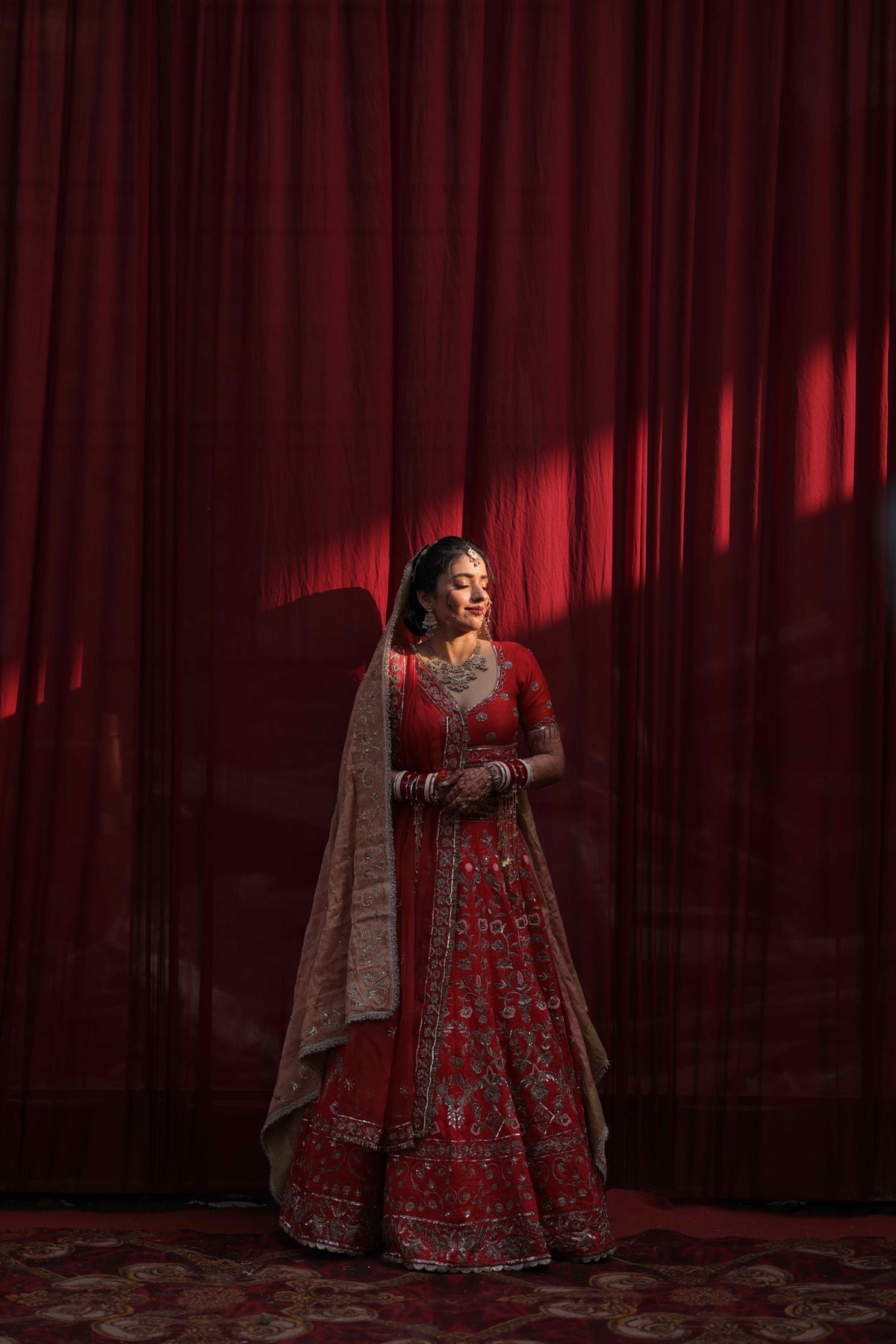 A woman wearing a red and gold embroidered traditional bridal lehenga with a peach dupatta stands in front of a dark red curtain backdrop with dramatic side lighting