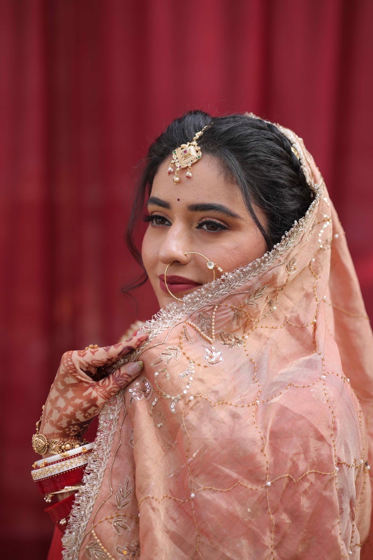 A woman wearing traditional Indian bridal attire, featuring a peach-colored embroidered dupatta, a red and gold lehenga, and ornate gold jewelry, posed against a draped red curtain background.