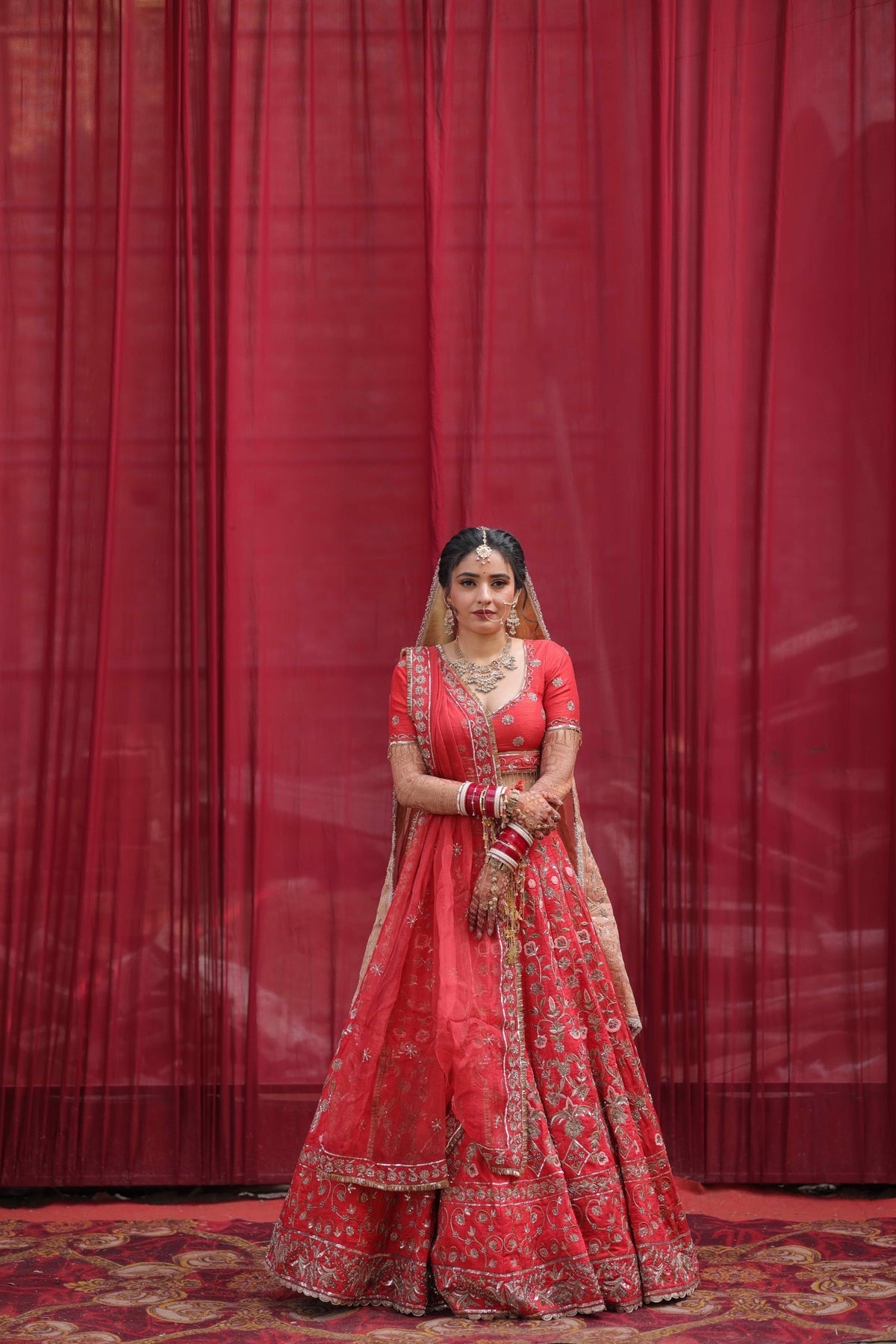 A woman wearing a traditional red and gold embroidered Indian lehenga choli outfit stands in front of a red curtain backdrop.