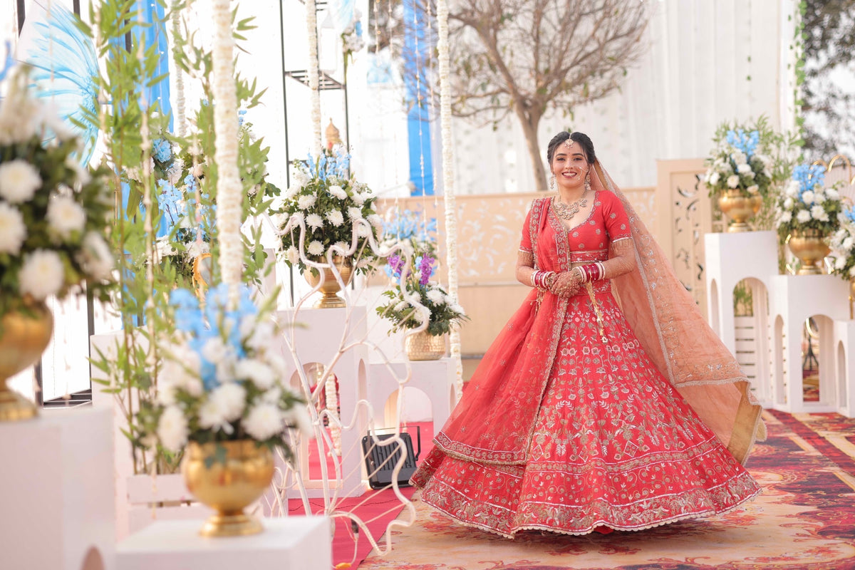 A woman wearing a bright red traditional bridal lehenga choli with intricate gold embroidery and a peach dupatta, standing in a decorated event hall with floral arrangements.