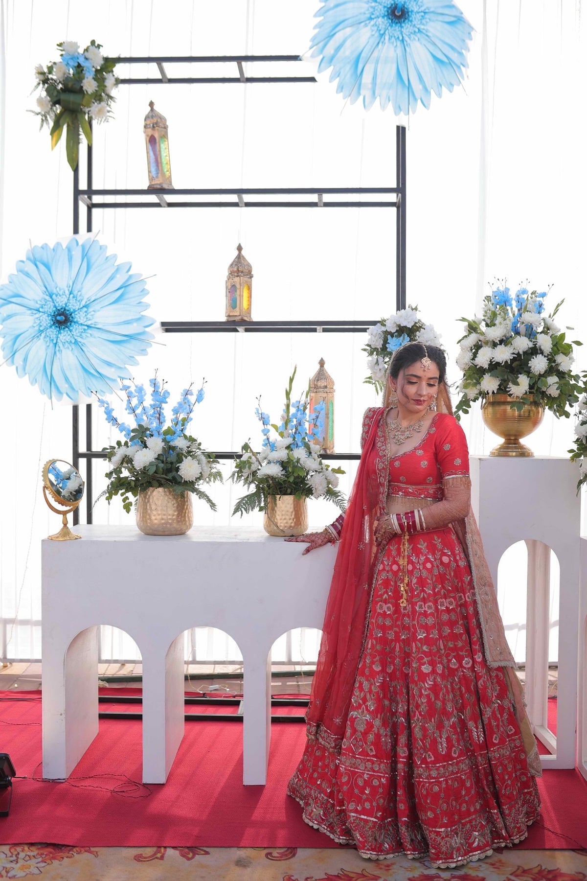 A woman wearing a red and gold heavily embroidered traditional Indian lehenga choli with a dupatta stands in front of a decorated white backdrop with floral arrangements and blue paper flowers.