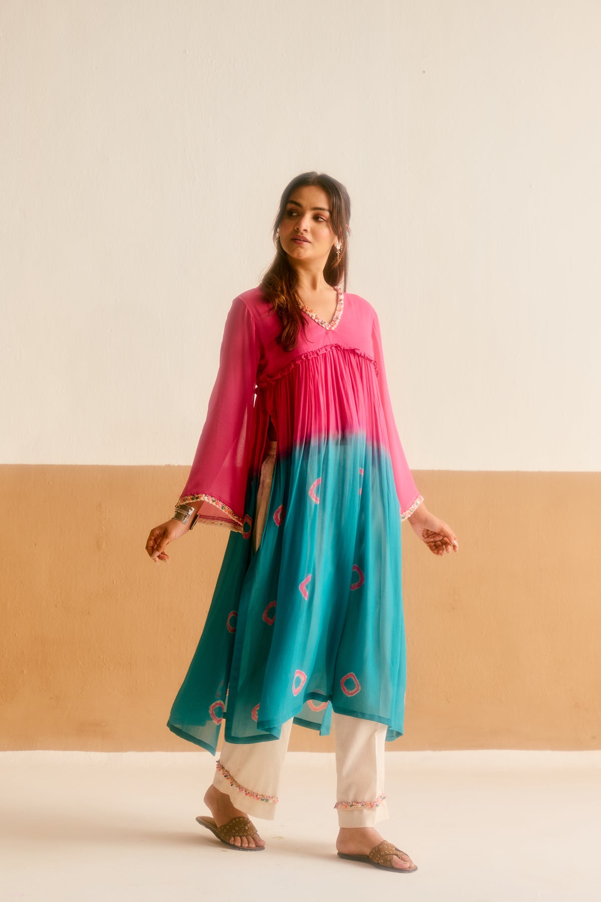 Woman wearing a pink and teal hand tied and dyed kurta with white pants against a beige and brown background