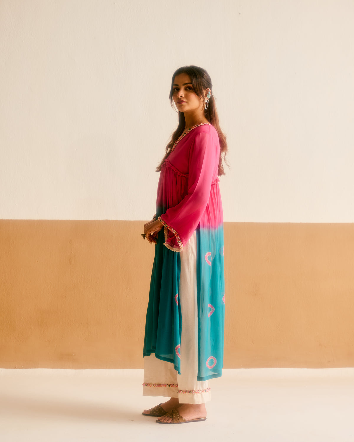A woman stands in profile wearing a traditional outfit with a bright pink and teal top featuring circular patterns, and cream pants, set against a minimalist beige and white wall background.