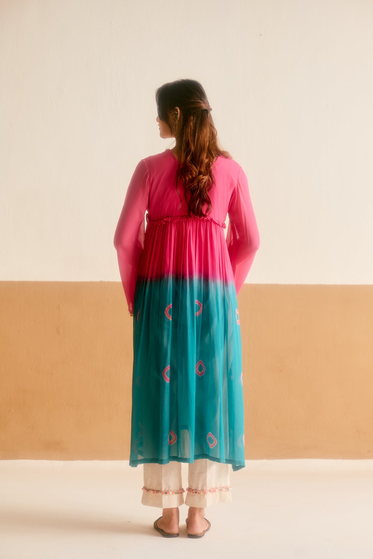 A woman viewed from behind wearing a long-sleeved pink and teal ombre tunic kurta with white pants, standing against a two-tone beige and off-white wall.
