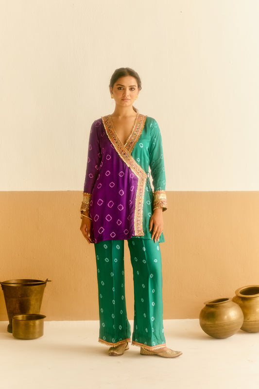 Woman wearing a green and purple hand tied and dyed outfit, standing against a beige wall with decorative pots.