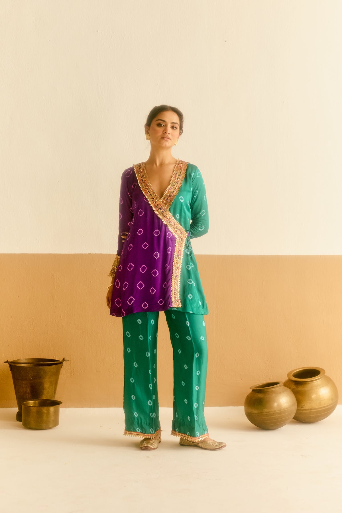A woman models a Indian ethnic suit consisting of a purple and green kurta with gold border, matching green pants, set against a beige and cream background with decorative brass pots.