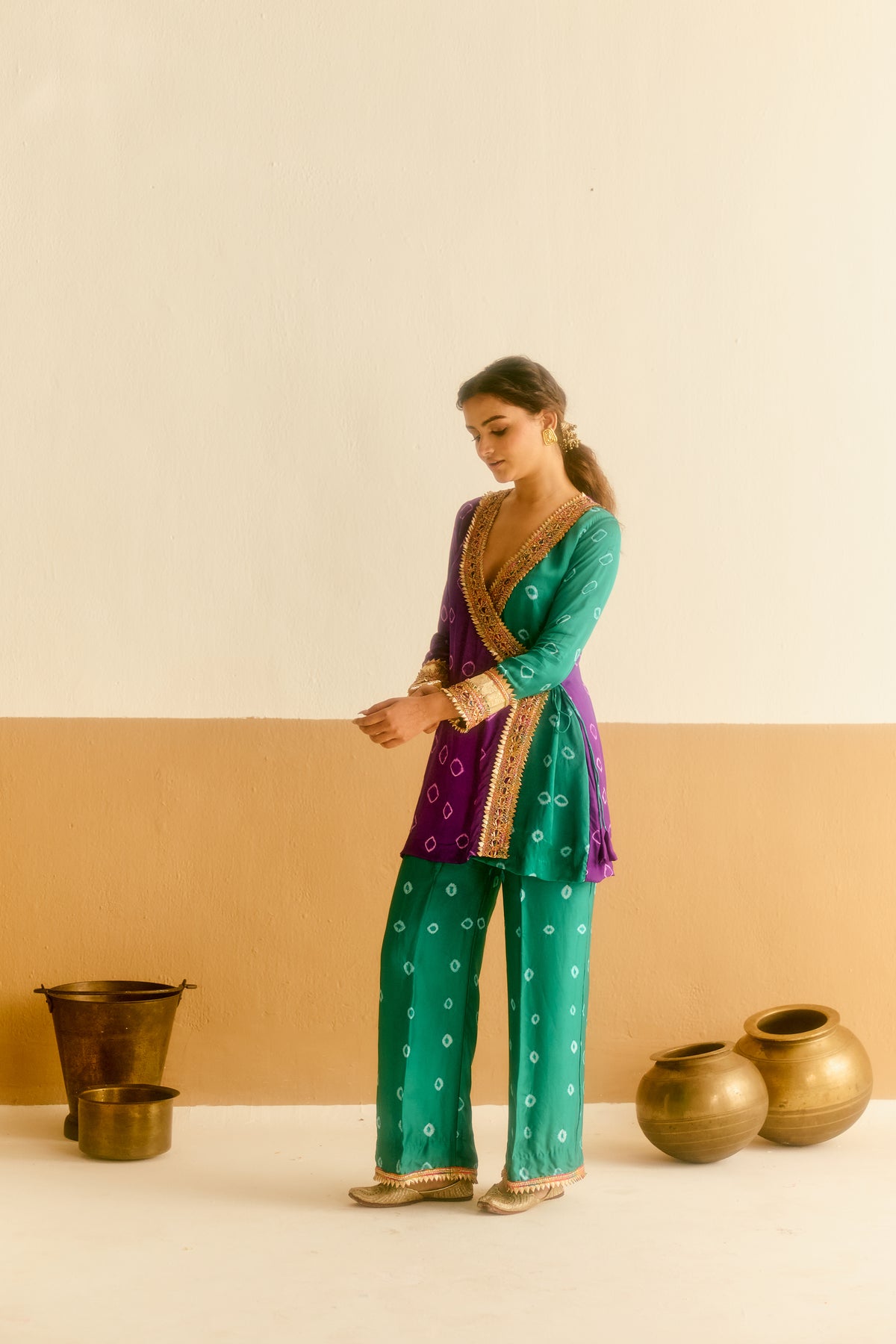 A woman modeling a bandhani angrakha and  salwar suit in green and purple with gold embroidery, standing in a studio with clay pots on the floor.
