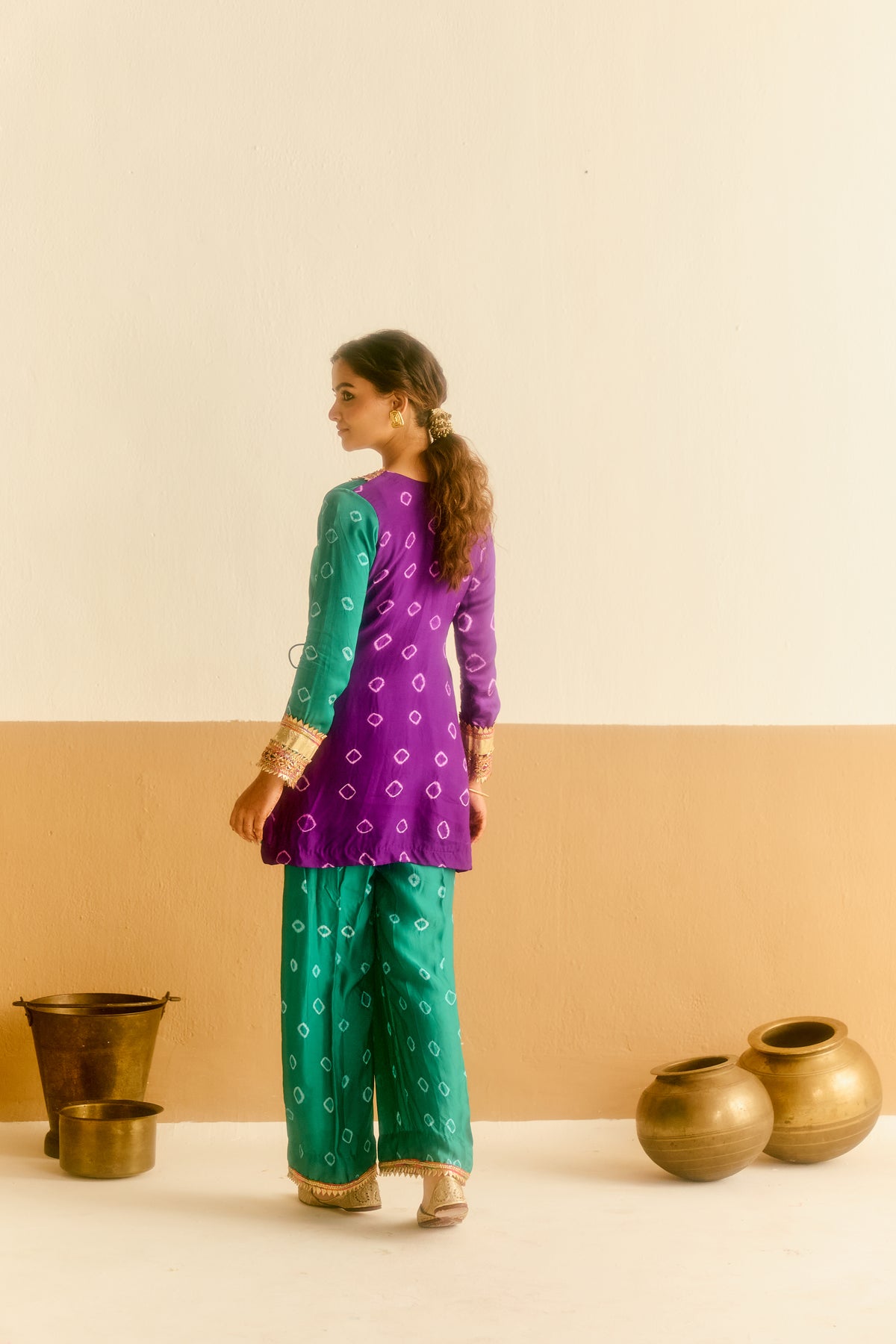Woman in a green and purple traditional outfit standing against a beige wall with decorative pots.