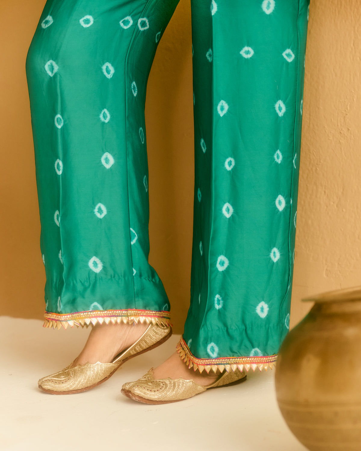 Green wide-leg pants with white tie-dye style diamond patterns and gold-trimmed hem, shown on a model wearing gold flat shoes against a warm beige wall.